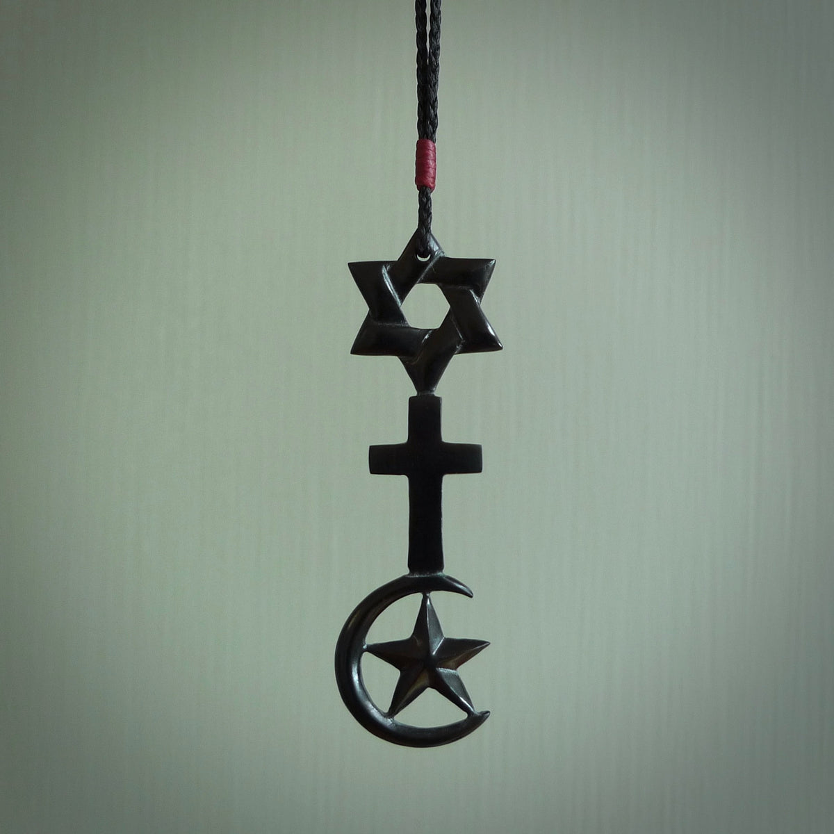 This pendant is a series of religious symbols carved in one piece of black jade. The top is a star of david, next is a christian cross and at the bottom is the crescent of islam. It is suspended on a fine, plaited black cord which is length adjustable.