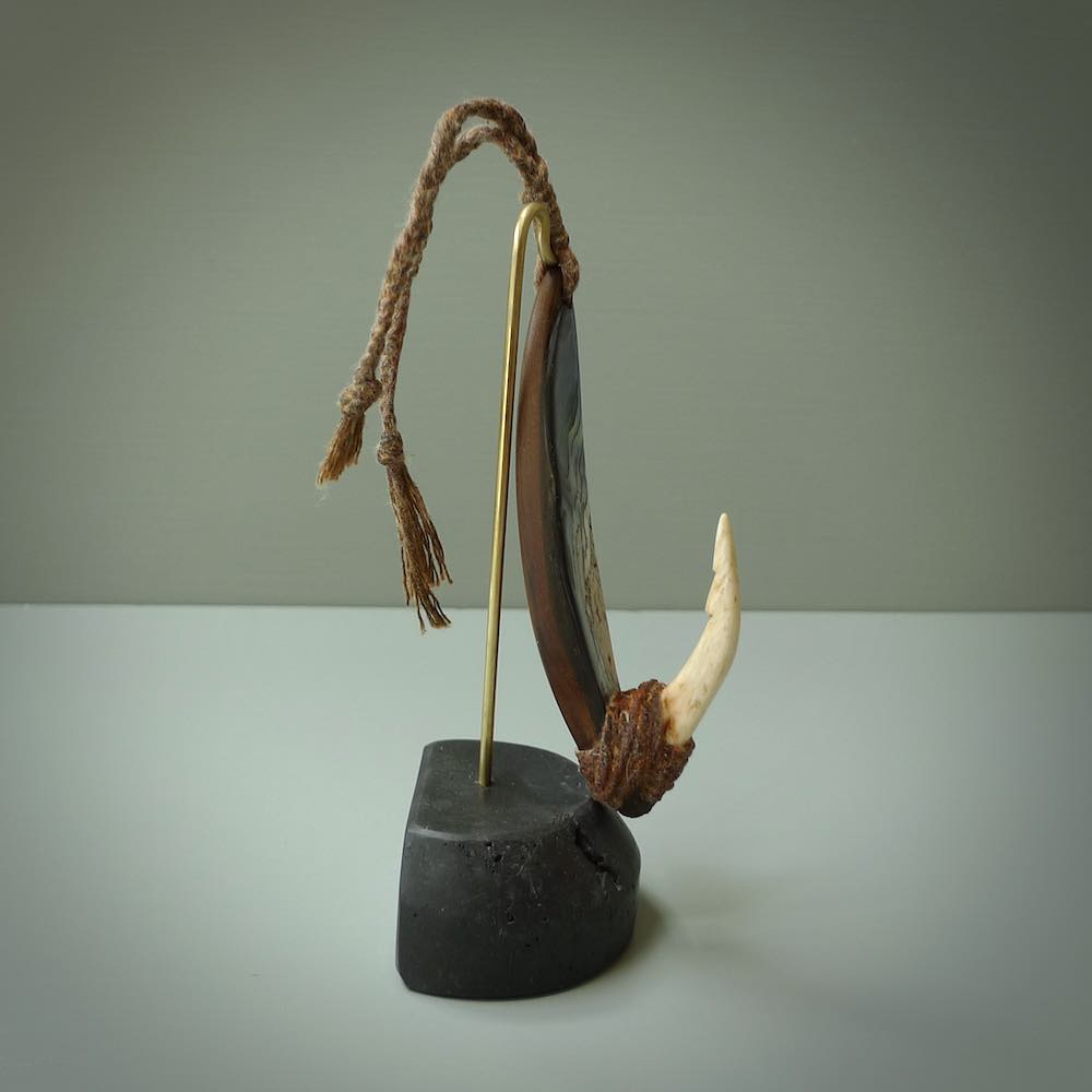 This picture shows a small sculptural hook called a pā kahawai. It is carved from whale bone, paua shell and wood, and is a colourful blue grey colour. It is shown on a stand made from argillite stone and is held in a brass cradle. One only, free shipping worldwide.