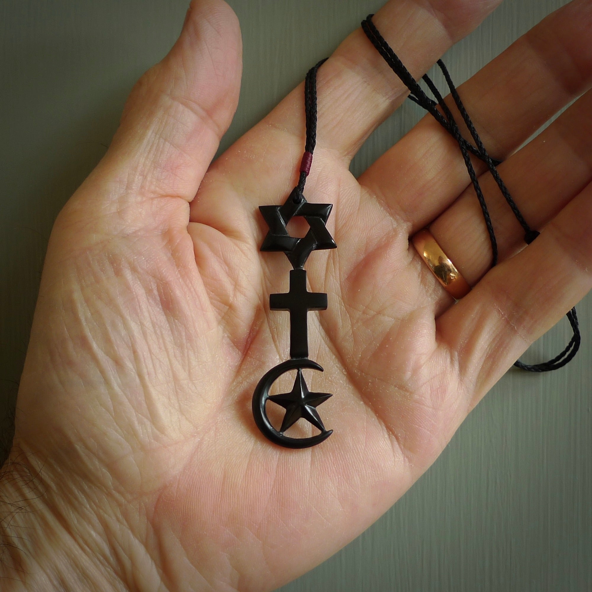 This pendant is a series of religious symbols carved in one piece of black jade. The top is a star of david, next is a christian cross and at the bottom is the crescent of islam. It is suspended on a fine, plaited black cord which is length adjustable.