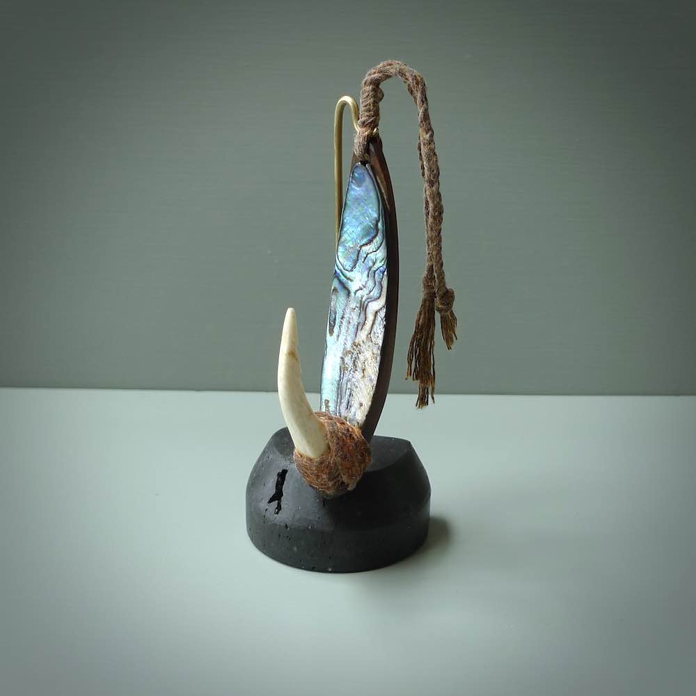 This picture shows a small sculptural hook called a pā kahawai. It is carved from whale bone, paua shell and wood, and is a colourful blue grey colour. It is shown on a stand made from argillite stone and is held in a brass cradle. One only, free shipping worldwide.