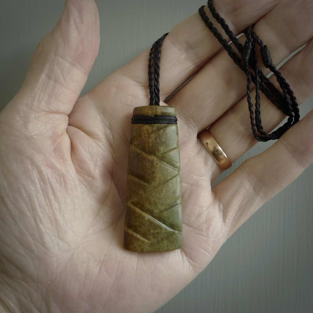 This picture shows a toki pendant carved with a woven front face and a flat back. It is made from a piece of rare New Zealand Flower Jade by our artist and carver Ana Krakosky. The cord is black and is length adjustable.