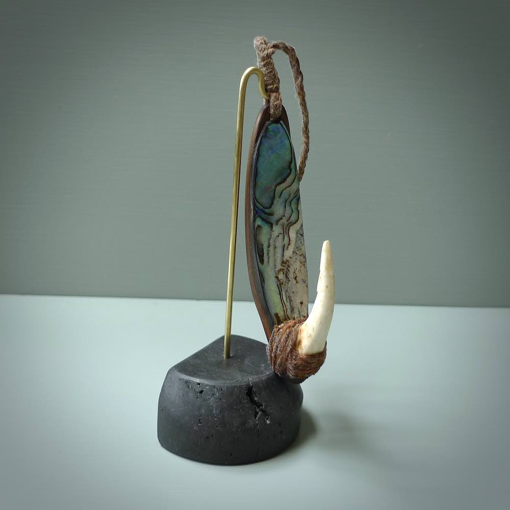 This picture shows a small sculptural hook called a pā kahawai. It is carved from whale bone, paua shell and wood, and is a colourful blue grey colour. It is shown on a stand made from argillite stone and is held in a brass cradle. One only, free shipping worldwide.